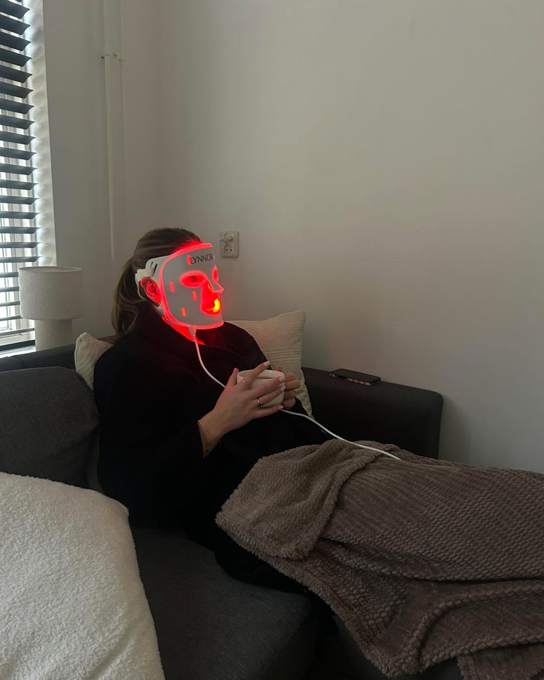 Persoon die een roodlichttherapie masker gebruikt in een rustige moderne woonkamer met zachte beige tinten.